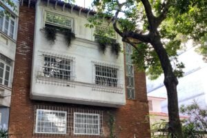 A imagem mostra uma casa de três andares, construída em tijolos, com uma fachada branca na parte superior. A casa possui janelas com grades e está cercada por um portão branco. Há vegetação ao redor, incluindo uma árvore à direita e plantas na base da casa. O ambiente é urbano, com calçada visível em primeiro plano.