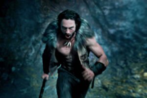 Um homem musculoso e sem camisa corre em um ambiente escuro e rochoso. Ele usa uma pele de animal nos ombros e tem um colar de dentes ou ossos. Sua expressão é intensa e determinada, e ele segura um bastão com uma das mãos.