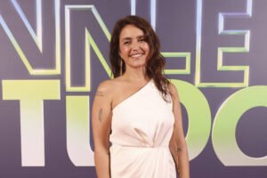 Uma mulher está posando em um evento de gala, vestindo um vestido longo branco com um ombro só. Ela sorri e está em pé sobre um tapete vermelho, com um fundo que apresenta o texto