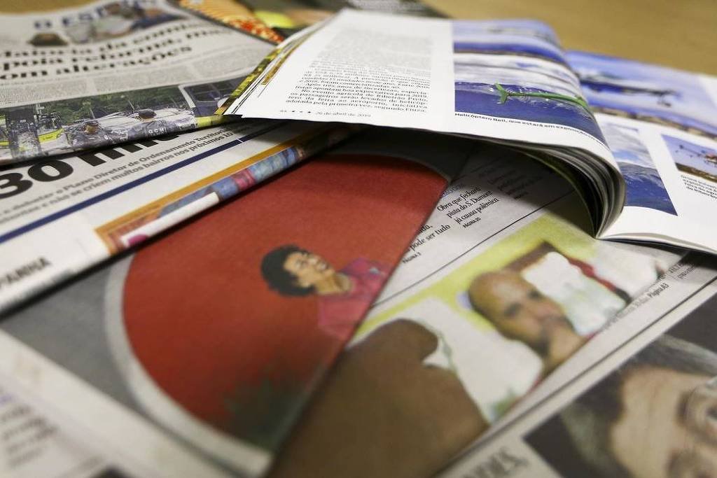 A imagem mostra uma pilha de jornais e revistas dispostos de forma desordenada sobre uma superfície. É possível ver partes de várias páginas, incluindo textos e imagens. Um dos jornais exibe a palavra