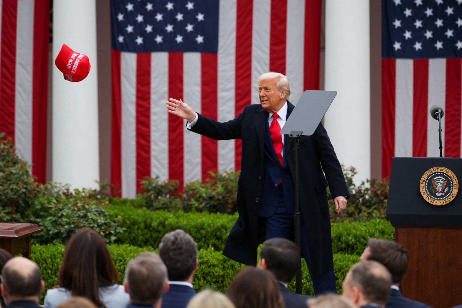 A imagem mostra um homem de terno escuro e gravata vermelha, em um evento ao ar livre, lançando um boné vermelho. Ao fundo, há bandeiras dos Estados Unidos e um público assistindo. O homem está em frente a um púlpito com o selo presidencial. A cena é iluminada e parece ser um momento de destaque durante o evento.
