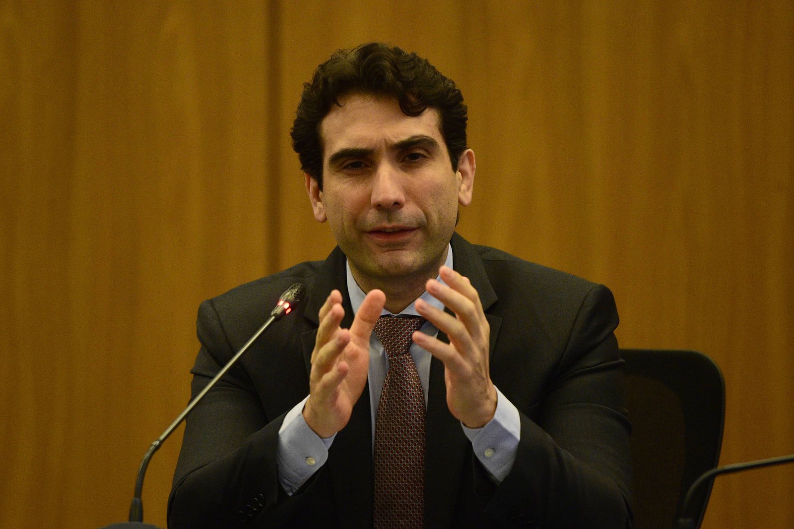 Um homem com cabelo escuro e encaracolado, vestindo um terno escuro e uma gravata, está sentado em uma mesa. Ele parece estar falando ou explicando algo, usando as mãos para gesticular. Ao fundo, há uma parede de madeira e um microfone na mesa à sua frente.