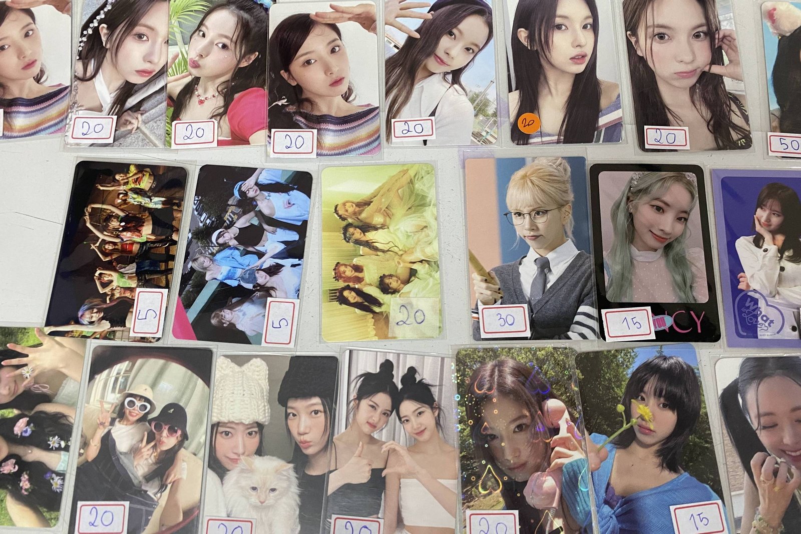 A imagem mostra uma coleção de cartões de fotos dispostos em uma superfície. Os cartões apresentam várias imagens de pessoas, possivelmente celebridades ou influenciadores, e estão organizados em linhas. Cada cartão tem um preço escrito à mão em um adesivo, variando entre 10 e 30. As fotos incluem retratos individuais e grupos, com diferentes expressões e estilos.