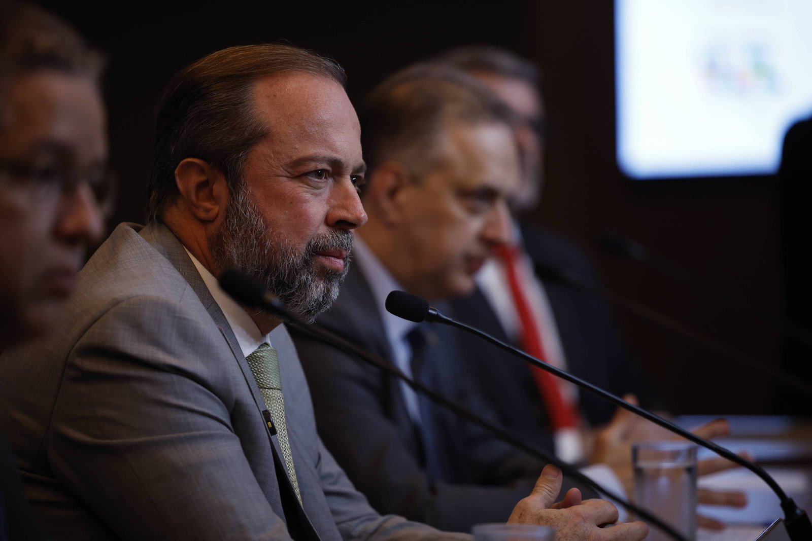 A imagem mostra um grupo de homens em uma mesa durante uma conferência. O homem em primeiro plano, com barba e cabelo castanho, está usando um terno cinza e uma gravata verde. Ele parece estar falando ou ouvindo atentamente. Ao fundo, outros homens estão visíveis, alguns com expressões sérias, e há um microfone na mesa. Uma tela ao fundo exibe um gráfico ou logotipo, mas não é possível ler o conteúdo.