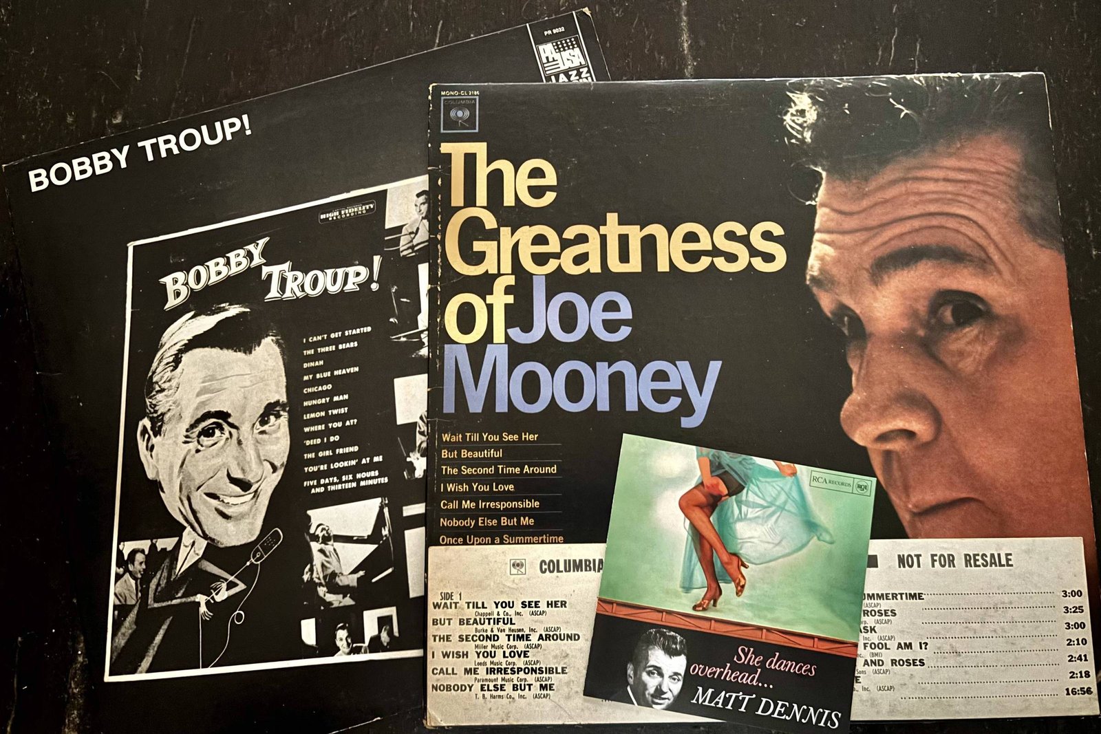 A imagem mostra três capas de discos de vinil sobre uma superfície escura. A capa superior é de