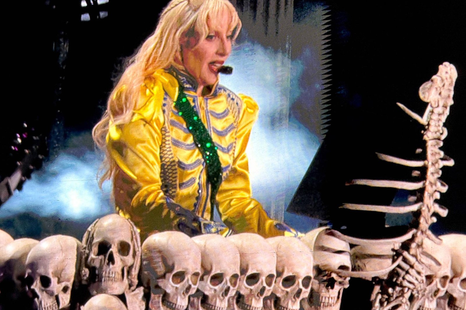 A imagem mostra uma performance de um artista em um palco. O artista está vestido com um traje amarelo brilhante e usa uma maquiagem marcante. Atrás dele, há uma estrutura decorativa composta por várias caveiras e uma coluna vertebral exposta. O fundo é iluminado com luzes e fumaça, criando uma atmosfera dramática.