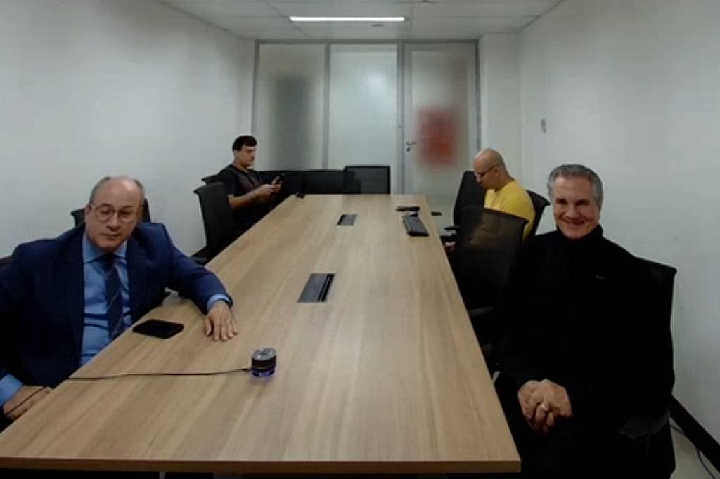 A imagem mostra uma sala de conferência com uma mesa longa e retangular. Há quatro homens sentados ao redor da mesa. Dois deles estão visíveis em primeiro plano: um homem à esquerda, com cabelo calvo e usando um terno azul, e outro à direita, com cabelo grisalho e usando uma camisa preta. Ao fundo, um homem está sentado em uma cadeira, usando uma camisa amarela, e outro homem está em pé, aparentemente usando um celular. A sala é iluminada e possui paredes brancas.