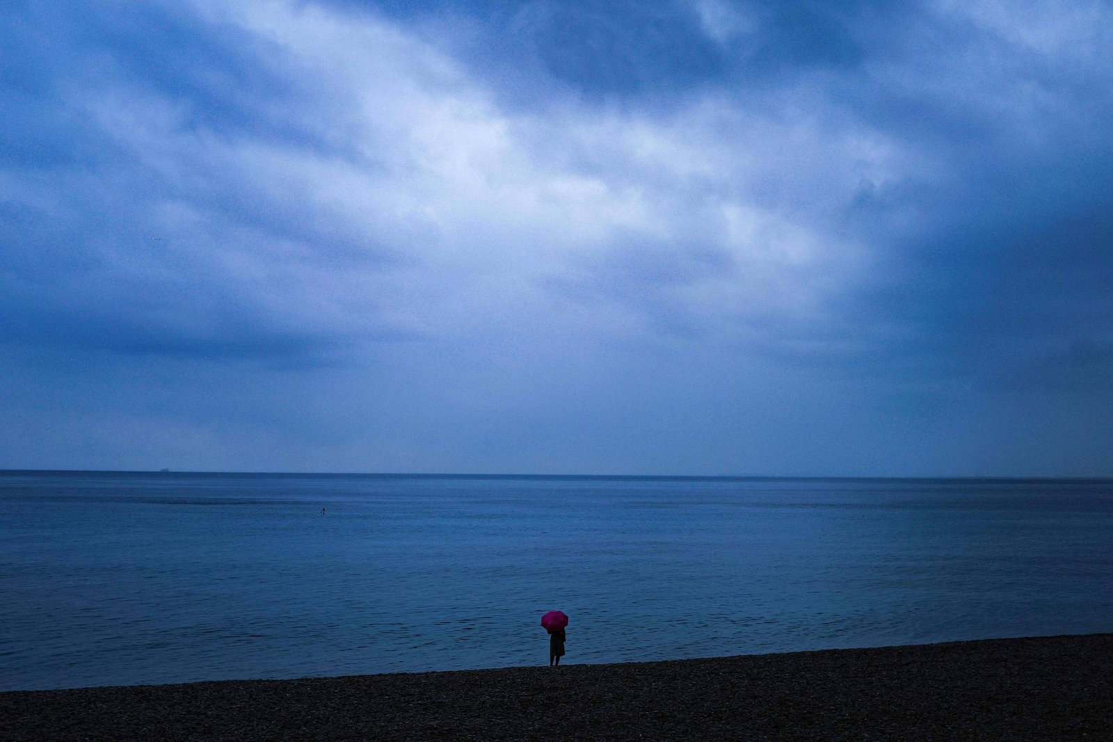 A imagem mostra uma pessoa sozinha na praia, segurando um guarda-chuva rosa. O céu está nublado, com tons de azul e cinza, refletindo uma atmosfera melancólica. O mar é calmo e se estende até o horizonte, onde a luz é mais clara. A areia da praia é visível na parte inferior da imagem.