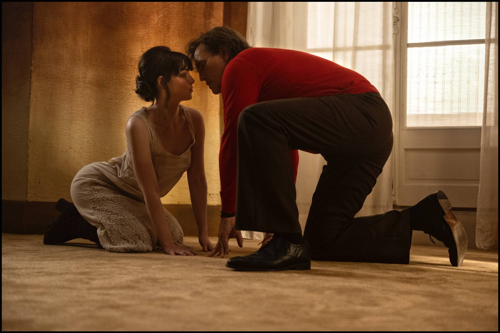 A imagem mostra um homem e uma mulher em uma posição íntima, ambos de joelhos no chão. A mulher está vestindo um vestido claro e está inclinada para frente, enquanto o homem, vestido com uma camisa vermelha e calças escuras, se aproxima dela. O ambiente é iluminado suavemente, com uma janela ao fundo e paredes de tom neutro.