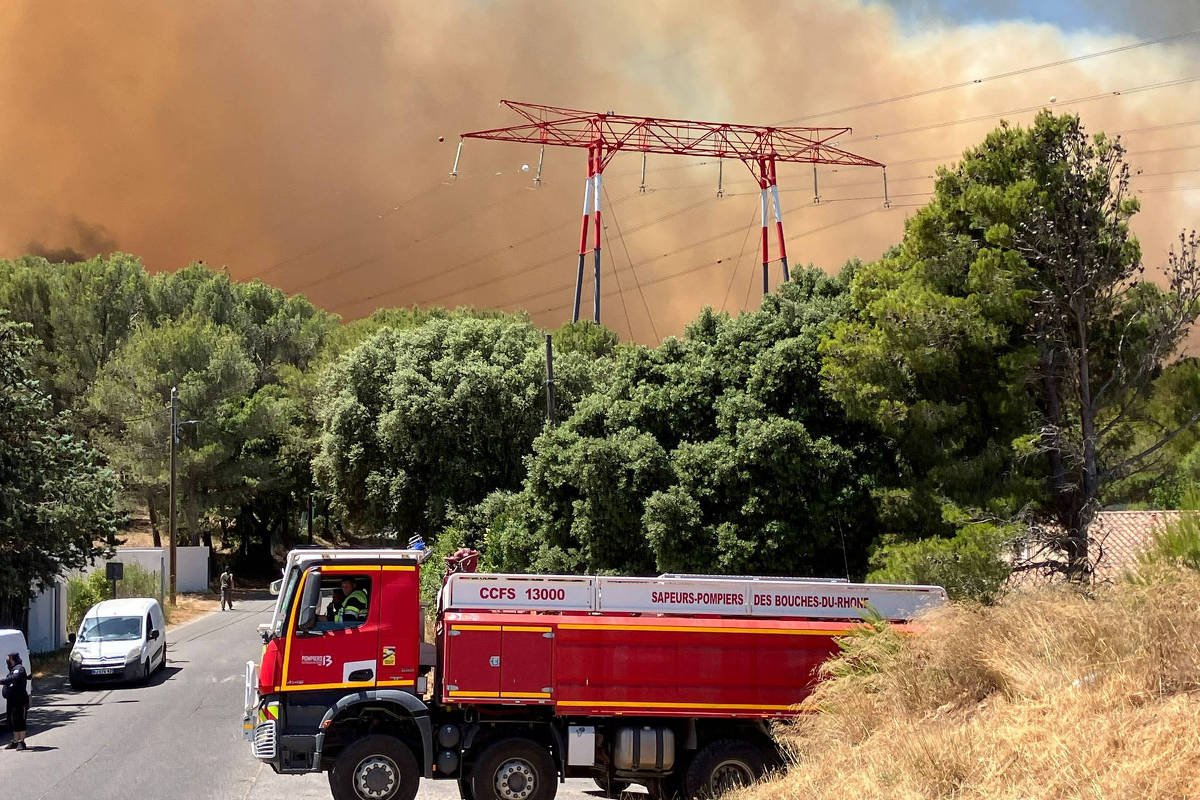 A imagem mostra um caminhão de bombeiros estacionado em uma rua, com vegetação densa ao redor. Ao fundo, há uma grande nuvem de fumaça laranja, indicando um incêndio florestal. Estruturas de energia elétrica são visíveis, com torres de transmissão se destacando contra o céu. O cenário é de emergência, com a presença de árvores e um terreno seco.