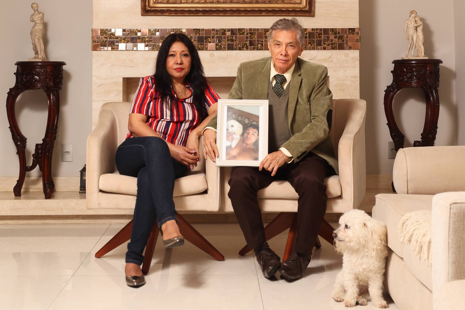 Um casal está sentado em sofás em uma sala de estar. A mulher, com cabelo longo e liso, usa uma blusa listrada em vermelho e azul e calças escuras. O homem, com cabelo grisalho, veste um paletó verde e segura uma moldura com uma foto. Ao lado deles, há um cachorro pequeno e branco. O ambiente é bem iluminado e decorado com estátuas e móveis de madeira.