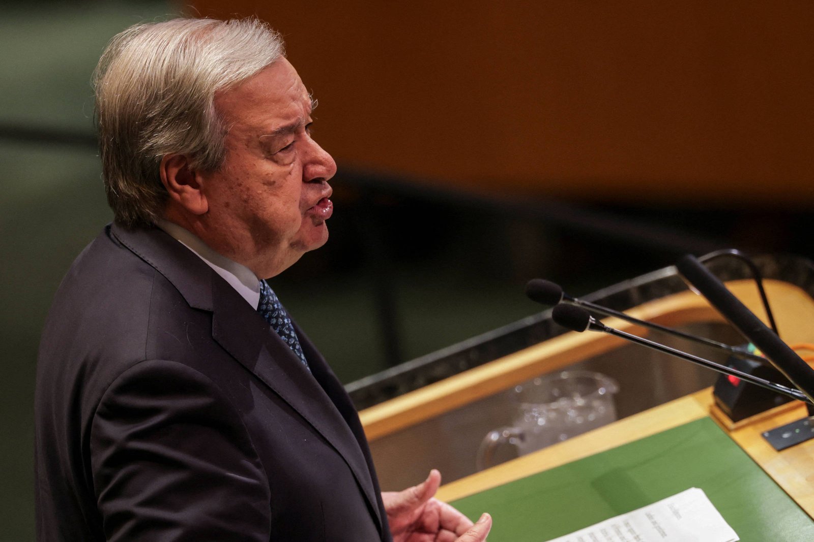 Um homem de cabelo grisalho e terno escuro está falando em um púlpito na Assembleia Geral da ONU. Ele está gesticulando com a mão esquerda enquanto segura um documento com a mão direita. Ao fundo, há um ambiente institucional com um painel e microfones.