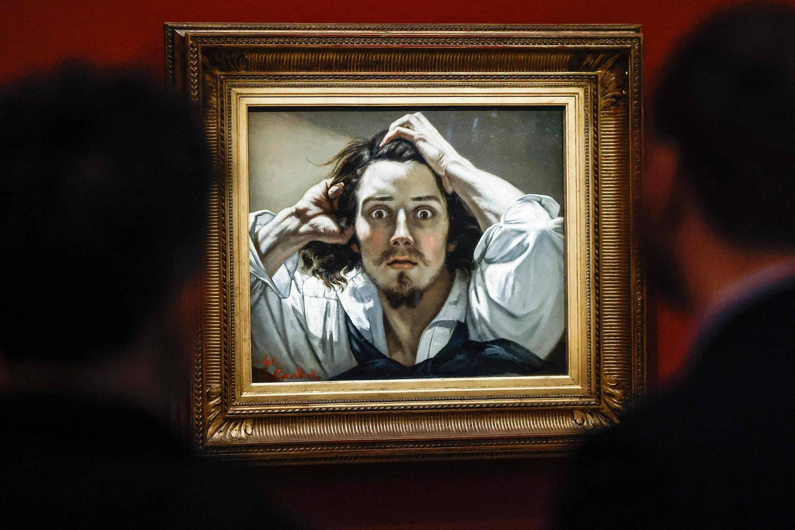 Dois homens observam pintura realista de homem de cabelos longos, camisa branca e colete preto, com expressão intensa e mãos na cabeça, em parede vermelha.