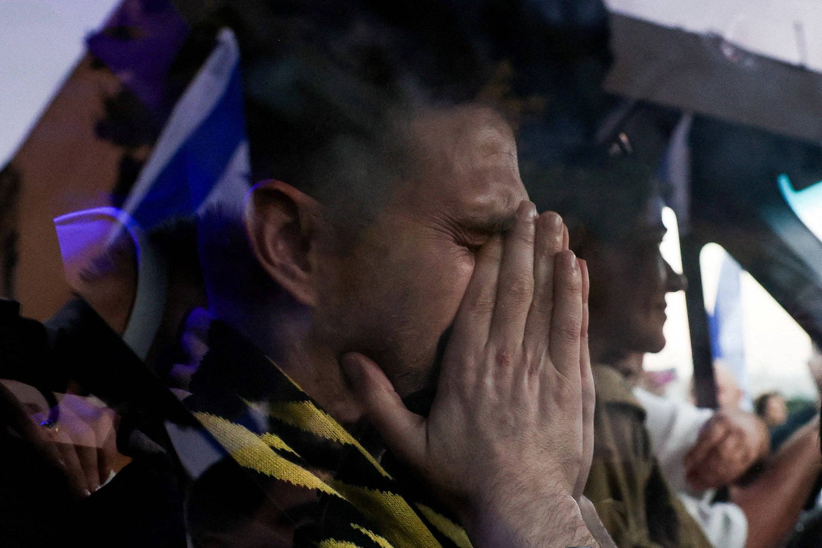 Homem com olhos fechados e mãos cobrindo o rosto sentado no banco dianteiro de um carro. Outra pessoa aparece ao fundo, também dentro do veículo, com expressão neutra.