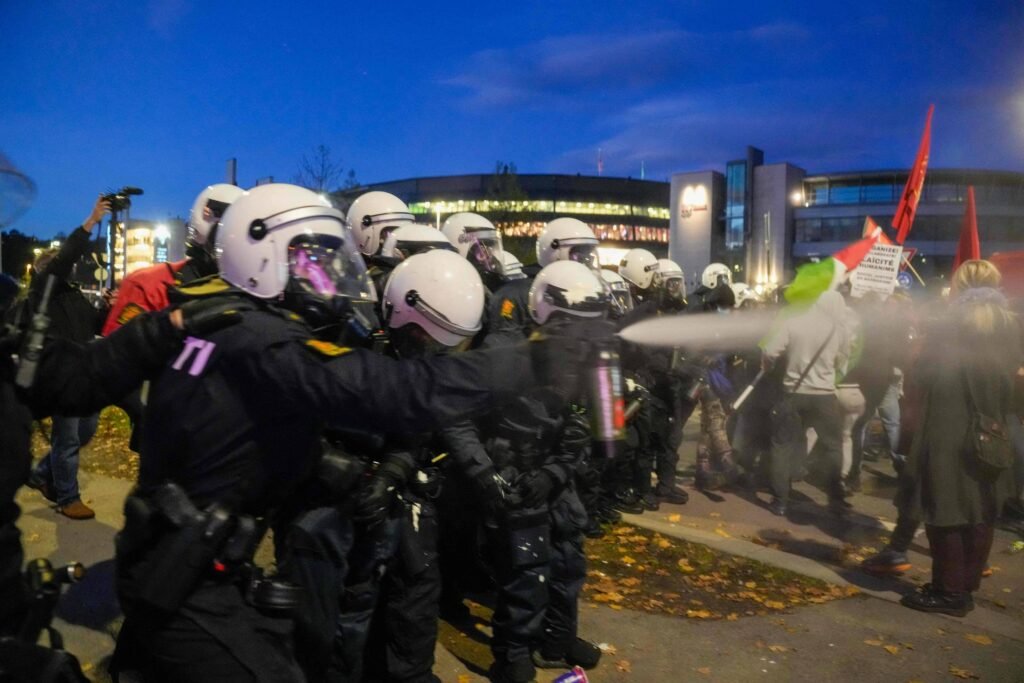 Policiais em linha, usando capacetes brancos e uniformes pretos, dispersam manifestantes com spray de pimenta em área externa urbana ao anoitecer. Manifestantes seguram bandeiras vermelhas e cartazes.