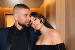 Homem de terno azul escuro com camisa preta e gravata borboleta abraça mulher com vestido preto de ombros à mostra e colar brilhante. Ambos estão próximos, em ambiente com paredes de madeira clara.