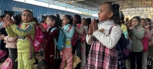 Um grupo de crianças palestinas está em pé em um centro de aprendizagem temporário em Gaza, aplaudindo, vestindo roupas coloridas e mochilas, com sinais do UNICEF e texto árabe visíveis no fundo.