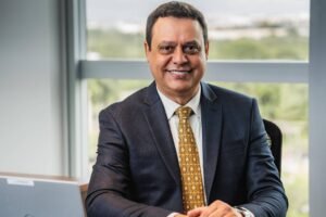 Um homem sorridente está sentado em uma mesa de escritório. Ele usa um terno escuro e uma gravata dourada. À sua frente, há um laptop prateado sobre a mesa. Ao fundo, uma grande janela permite a entrada de luz natural, mostrando uma vista de árvores e céu.