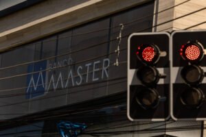 Semáforo com luz vermelha acesa em primeiro plano, com fachada do Banco Master ao fundo. Fios elétricos cruzam a imagem horizontalmente.