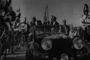 Carro conversível antigo com várias pessoas sentadas e em pé, cercado por cavaleiros fardados montados a cavalo. Bandeiras e multidão ao fundo em dia claro.