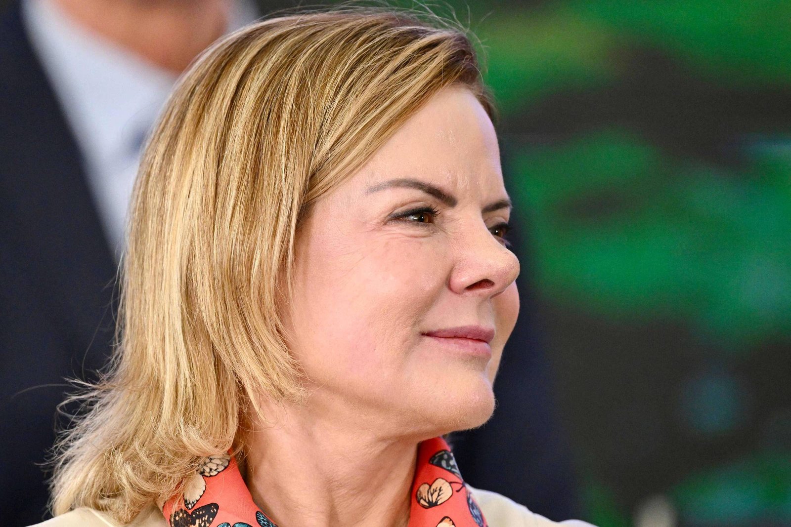 Mulher de cabelos loiros curtos, vista de perfil à direita, veste blusa clara e lenço laranja com estampa floral. Fundo desfocado com pessoa e vegetação.