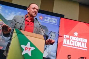 Homem de meia-idade com blazer cinza e camisa vermelha fala em púlpito com microfone. Fundo exibe telas com logo do PT e texto