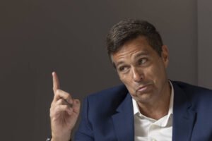 Um homem com cabelo castanho e curto, vestindo um paletó azul e uma camisa branca, está sentado e gesticulando com a mão direita levantada, como se estivesse enfatizando um ponto. Ele tem uma expressão facial que sugere que está explicando ou discutindo algo. O fundo é de cor neutra, sem muitos detalhes.