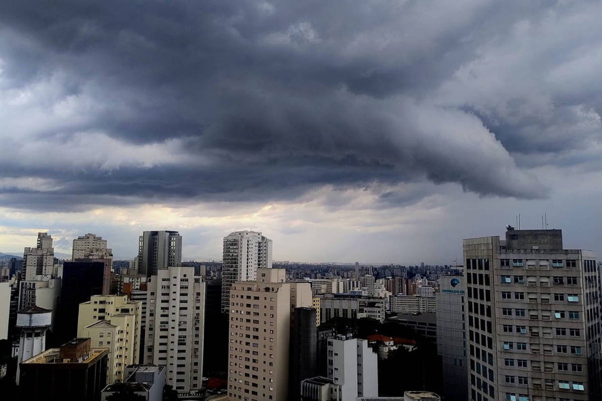 Nuvens escuras e densas cobrem o céu acima de prédios altos em área urbana, indicando tempestade iminente. Edifícios variados compõem o horizonte sob luz difusa.