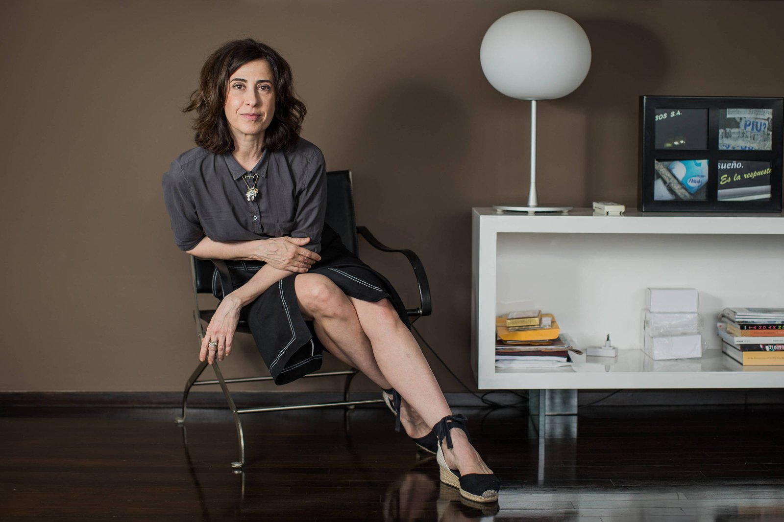 Mulher de cabelos castanhos ondulados sentada em cadeira preta com estrutura metálica, vestindo blusa cinza e saia preta, com pernas cruzadas. Ao lado, móvel branco com livros, objetos decorativos e luminária de mesa branca. Fundo marrom e piso escuro refletem luz natural de janelas ao fundo.