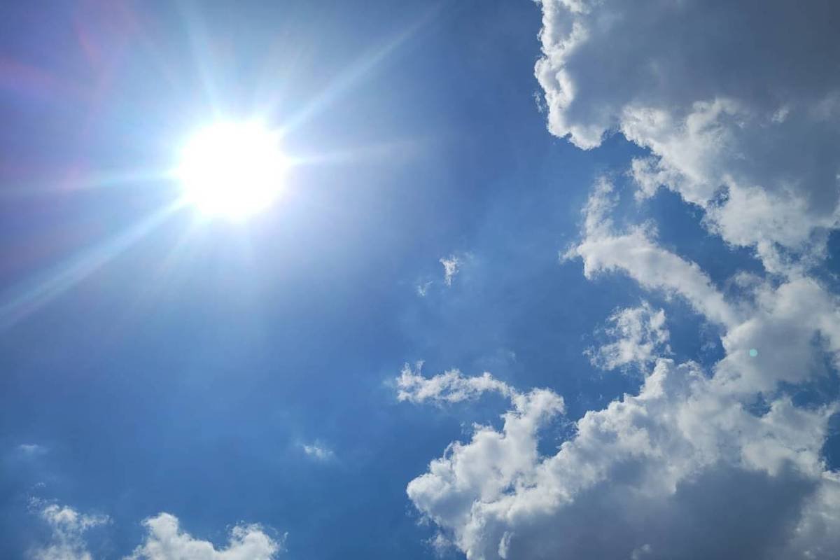 Sol intenso no canto superior esquerdo com raios visíveis, céu azul claro e nuvens brancas dispersas principalmente à direita e na parte inferior da imagem.