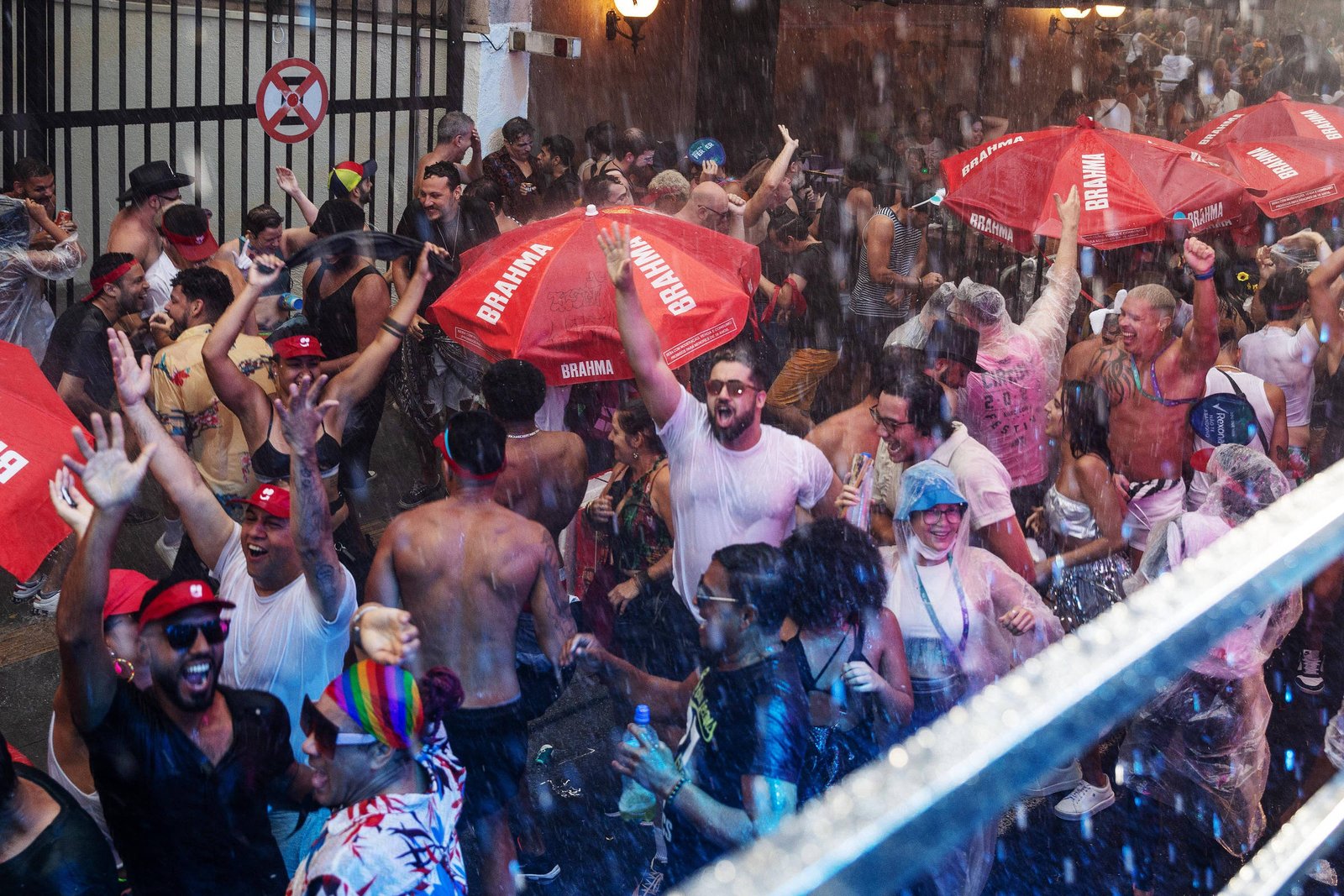 Multidão de pessoas celebra em festa ao ar livre sob chuva intensa. Alguns seguram guarda-sóis vermelhos com a marca Brahma, enquanto outros dançam e levantam os braços. Ambiente animado e lotado, com iluminação artificial e roupas variadas.