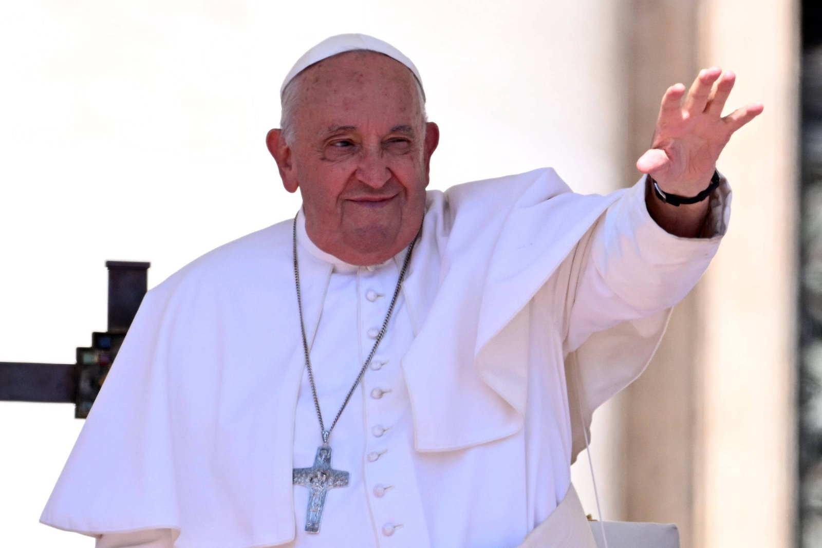 Um homem idoso, vestido com uma batina branca e um manto, está acenando com a mão direita. Ele usa um crucifixo pendurado no pescoço e tem uma expressão amigável. O fundo é desfocado, sugerindo um ambiente ao ar livre.