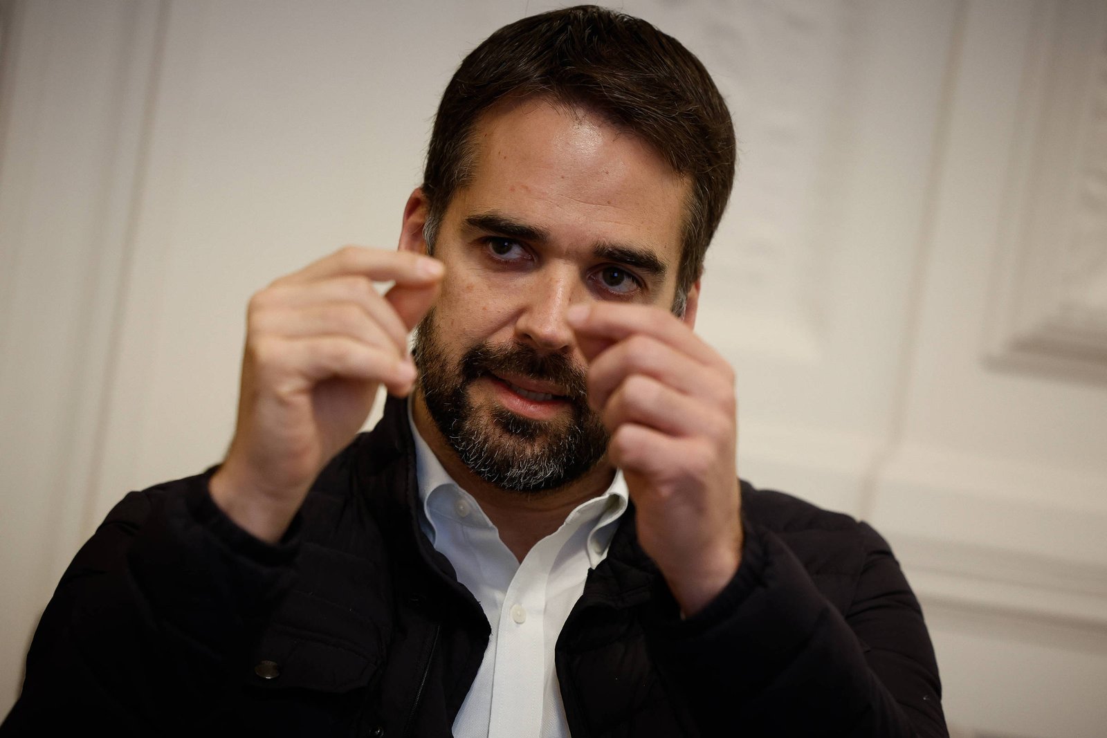 Homem de barba e cabelo curto, vestindo camisa branca e jaqueta preta, gesticula com as mãos próximas ao rosto em ambiente interno com parede clara ao fundo.