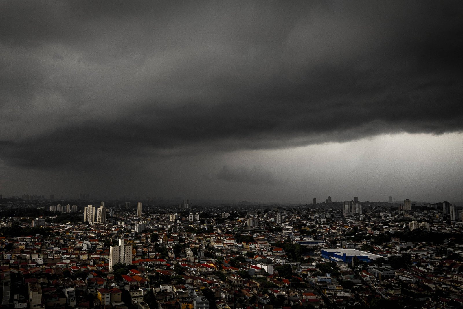 Nuvens escuras e densas cobrem o céu acima de uma grande cidade com prédios baixos e altos. A luz do sol ilumina parcialmente algumas áreas, enquanto outras permanecem na sombra sob a tempestade iminente.