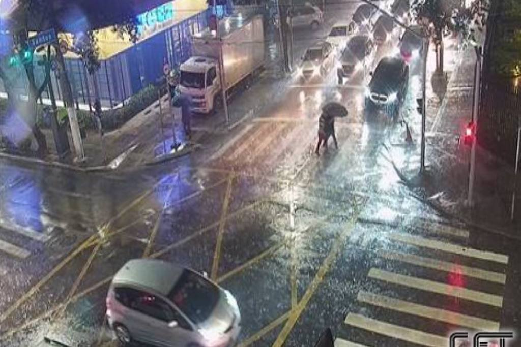 Cruzamento urbano à noite sob chuva forte, com ruas molhadas refletindo luzes. Dois pedestres atravessam a faixa com guarda-chuva aberto. Vários carros aguardam sinal vermelho, incluindo um caminhão estacionado próximo a um prédio iluminado.