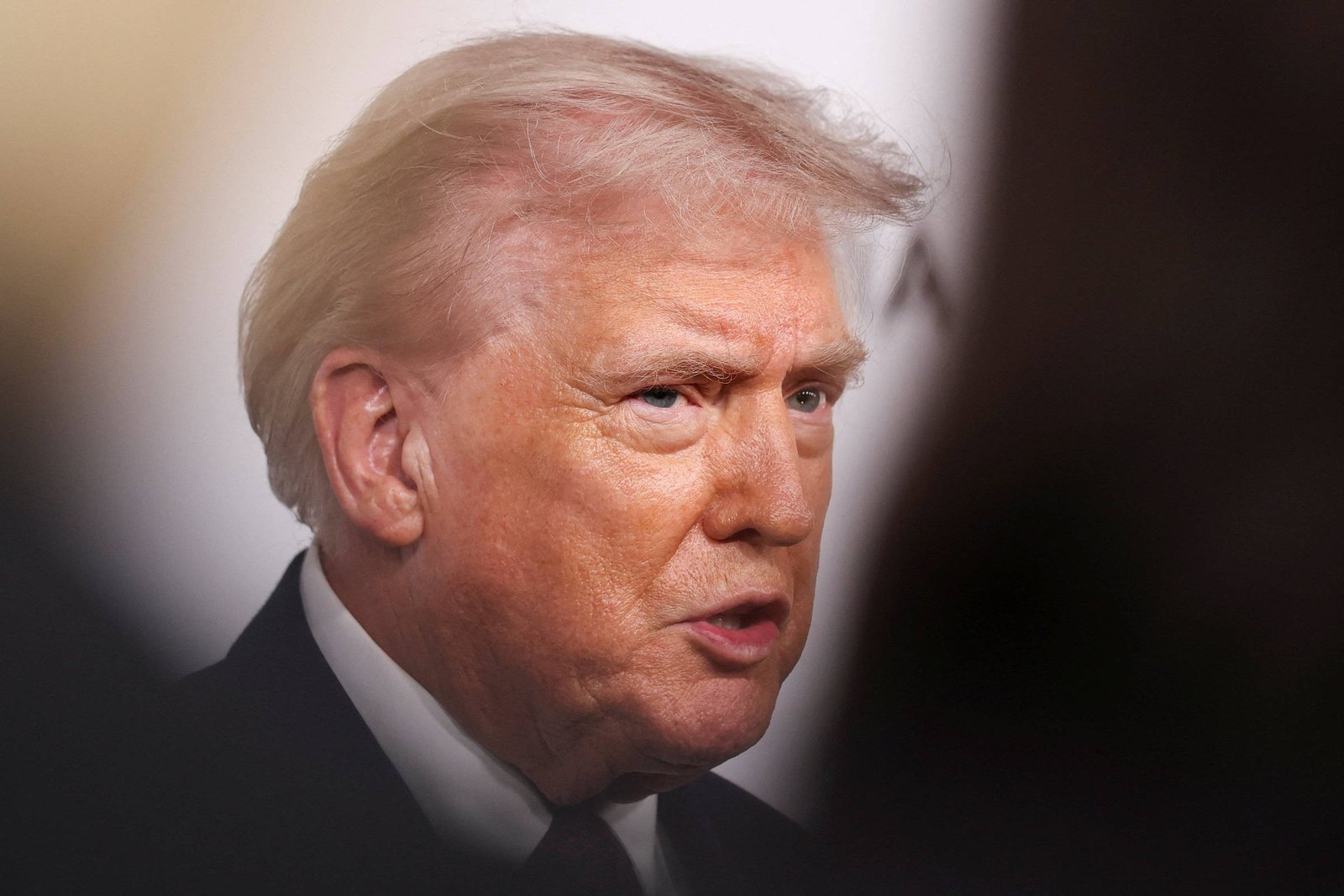 Homem branco de cabelo loiro esbranquiçado visto de perfil, com expressão séria, vestindo terno escuro e camisa branca. Fundo desfocado com áreas claras e escuras.