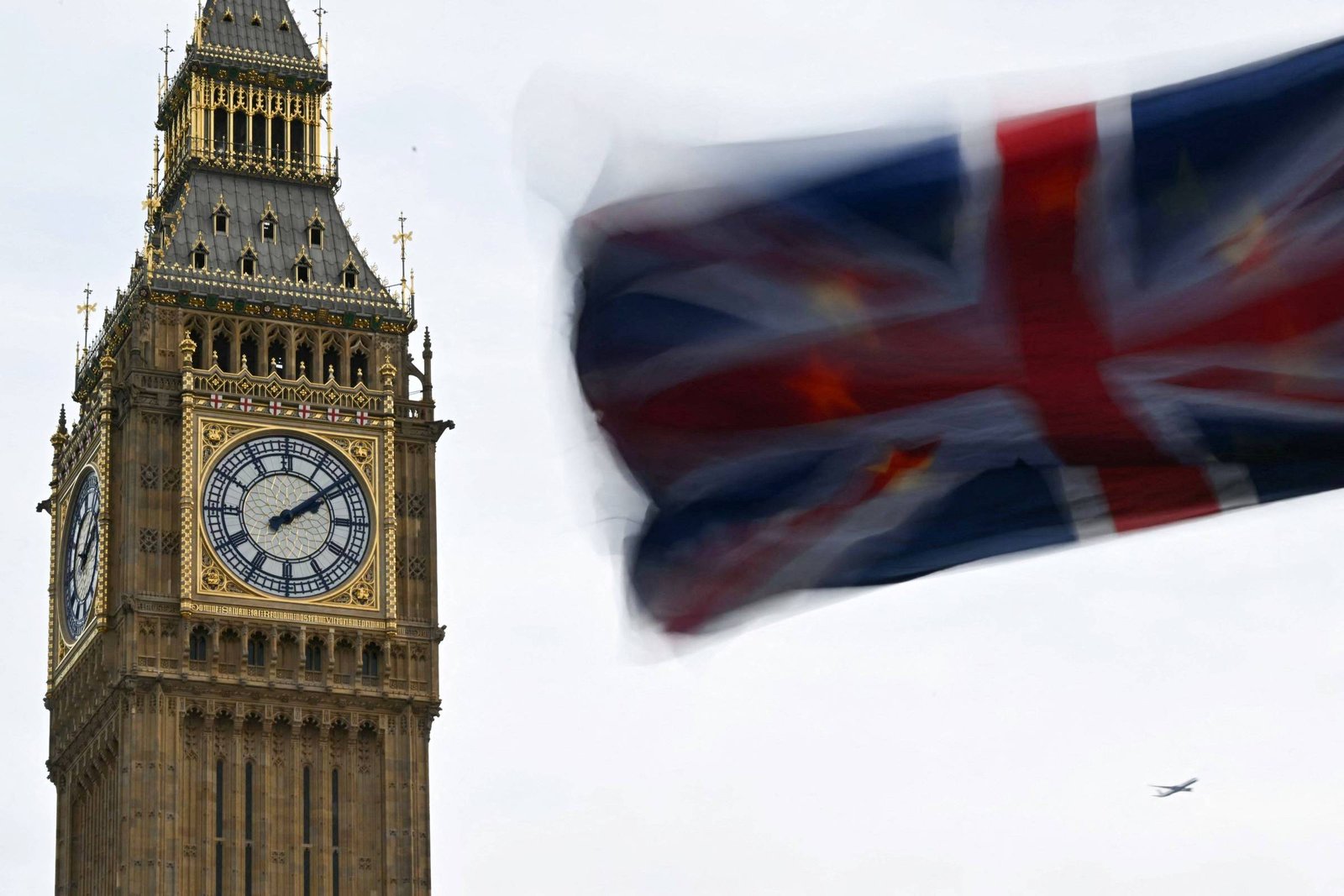 A imagem mostra a famosa torre do Big Ben, com seu grande relógio visível. Em primeiro plano, há uma bandeira do Reino Unido tremulando, que aparece um pouco desfocada. O céu está nublado, e a torre é predominantemente de cor dourada e cinza.