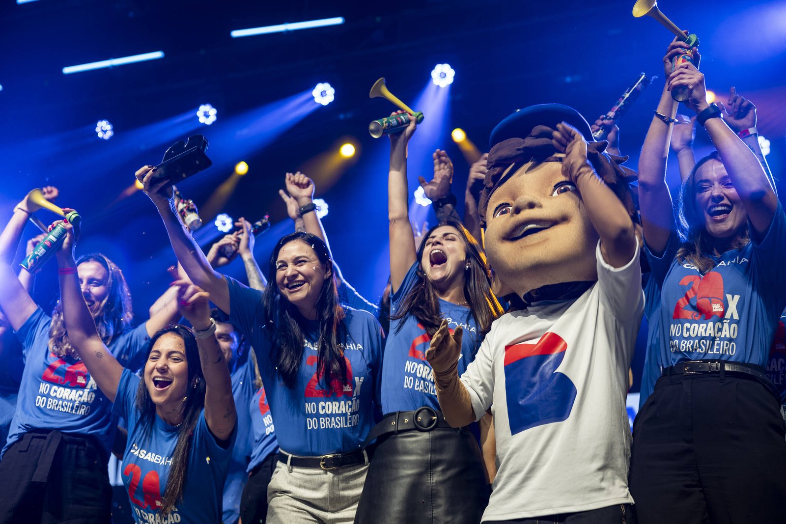 Pessoas vestindo camisetas azuis comemoram em palco iluminado, algumas segurando instrumentos de sopro e uma pessoa com fantasia de cabeça grande ao centro.