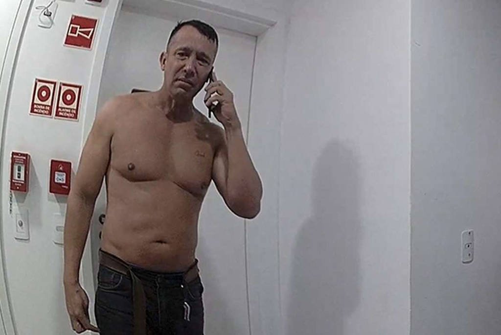 Homem sem camisa fala ao telefone em corredor estreito de prédio residencial. Outras pessoas estão ao fundo, algumas encostadas na parede, em ambiente interno com paredes brancas e portas ao lado.