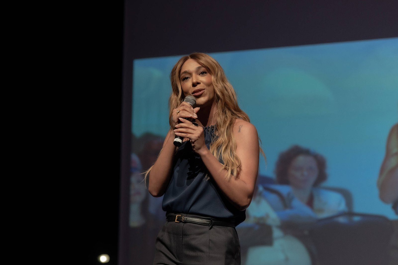 Uma mulher com cabelo longo e liso, usando uma blusa azul e calças escuras, está em um palco segurando um microfone. Ela parece estar falando para a audiência. Ao fundo, há uma projeção de uma apresentação com várias pessoas sentadas em uma mesa.