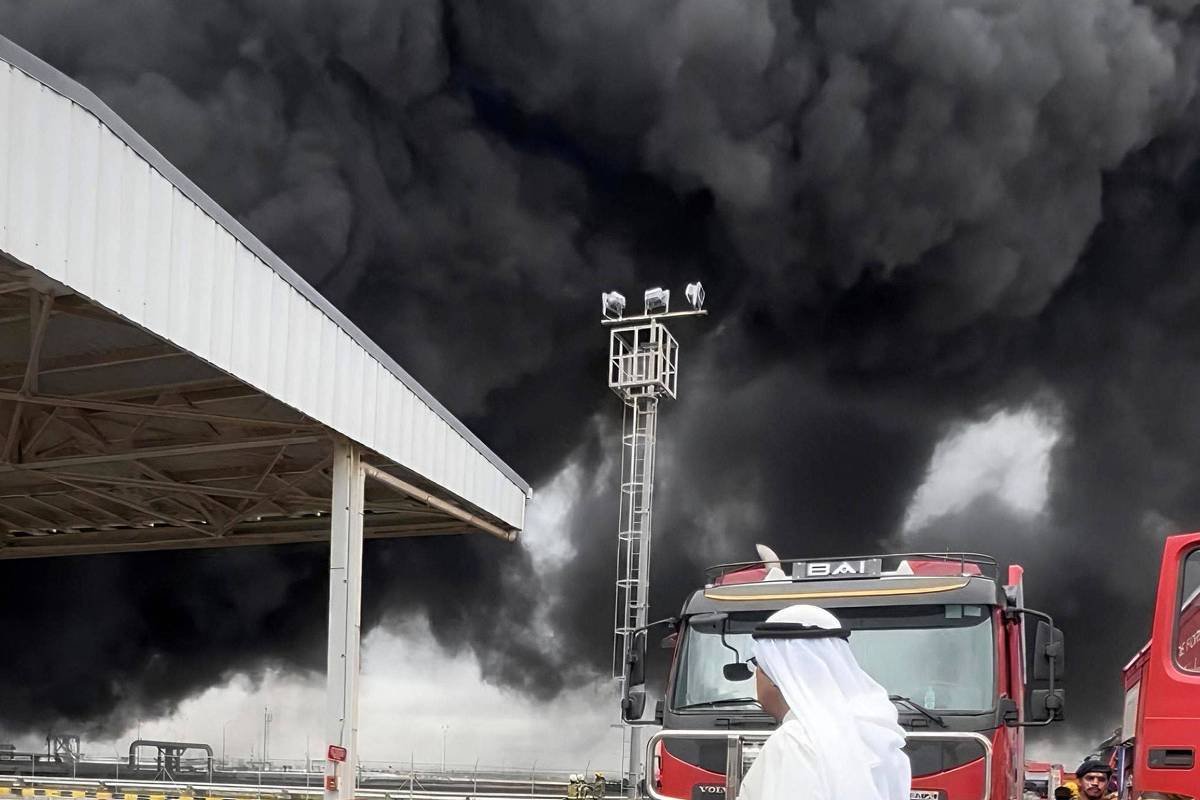 Nuvem espessa de fumaça preta cobre o céu sobre área industrial. Caminhões de bombeiros e bombeiros estão no local, com um deles em escada elevatória combatendo o fogo. Homem de roupa branca observa a cena em primeiro plano.