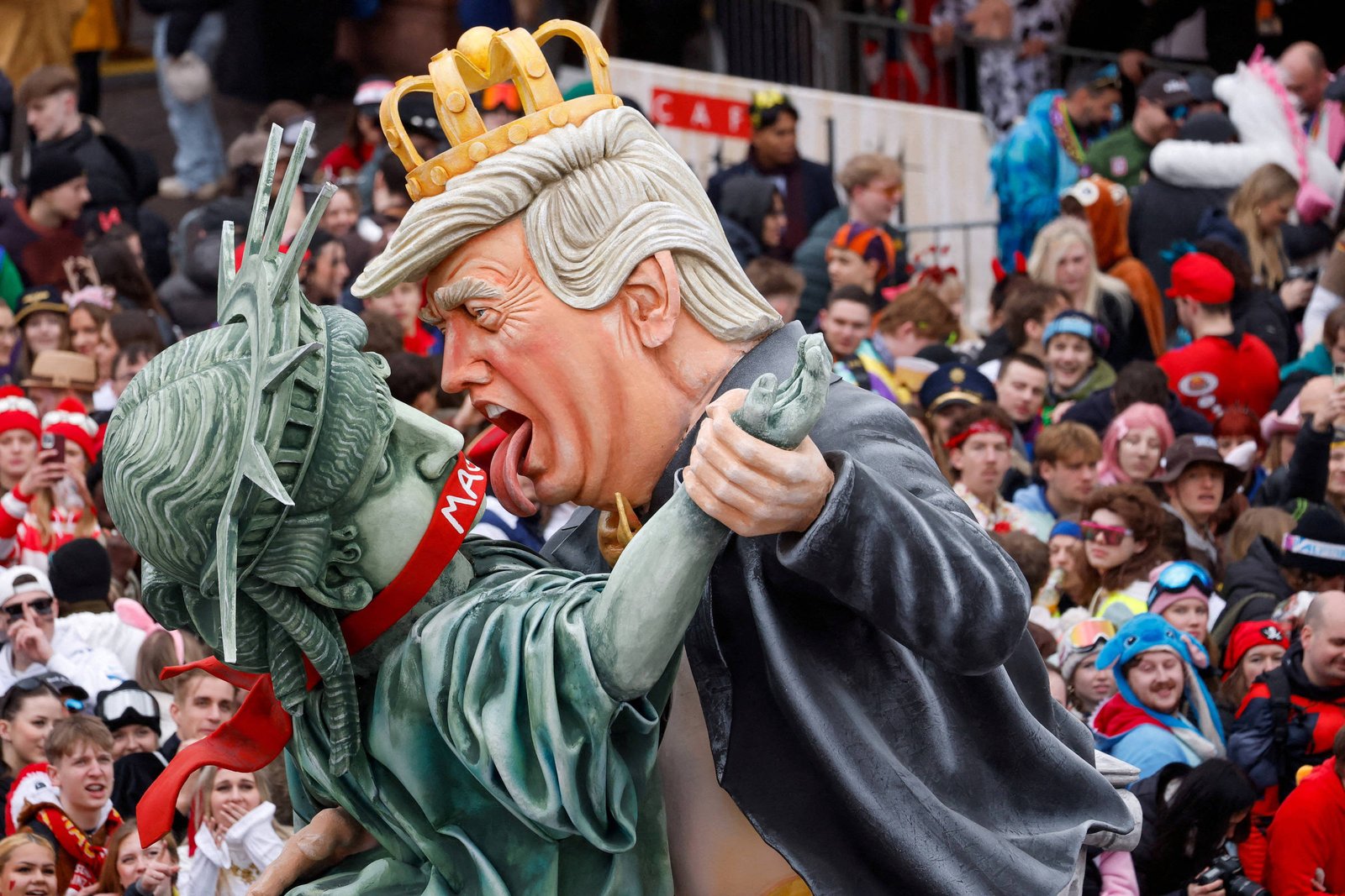 Bonecos gigantes caricaturais de Donald Trump com coroa dourada e da Estátua da Liberdade em pose de dança, cercados por multidão em evento ao ar livre.