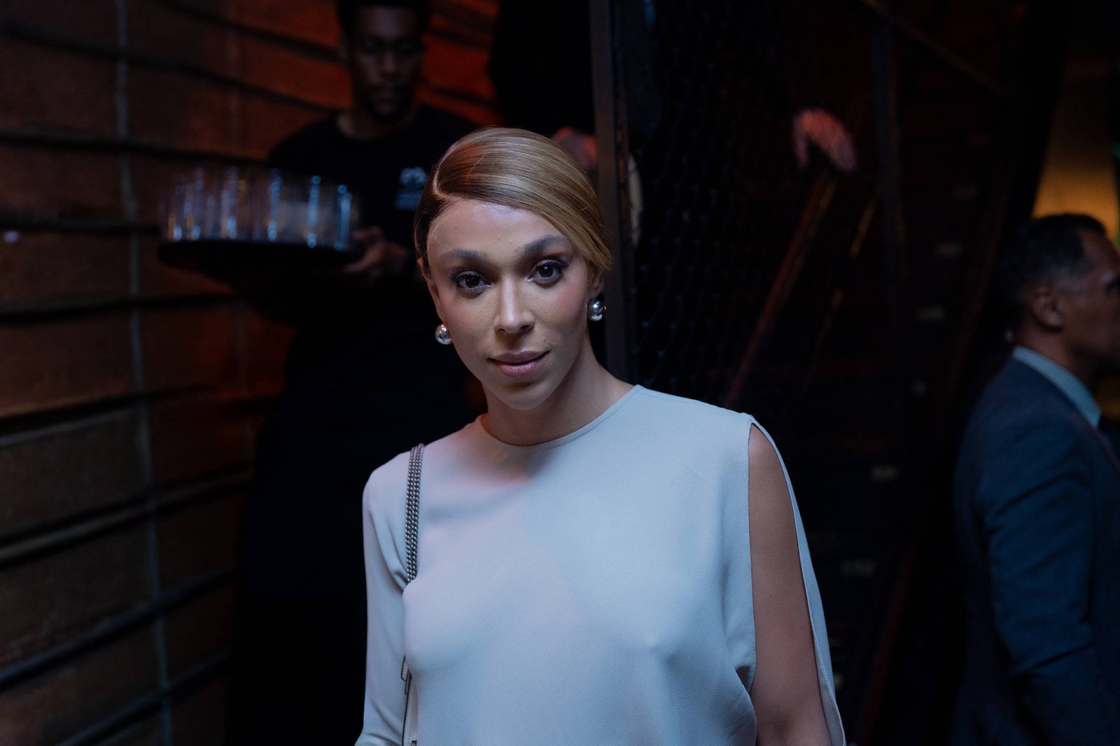 Pessoa com cabelo loiro preso, usando vestido branco sem mangas, posando em ambiente interno com iluminação baixa. Ao fundo, duas pessoas, uma carregando bandeja com copos e outra de terno azul.