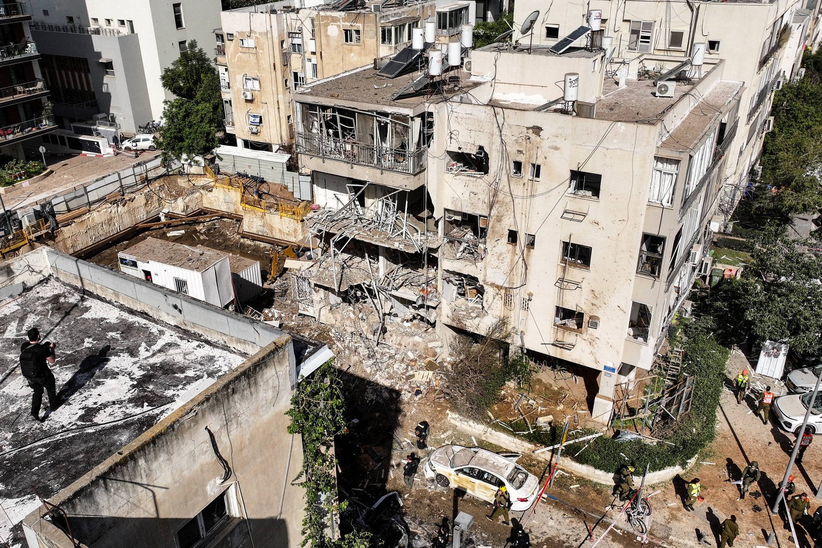 Prédio de quatro andares com fachada danificada e escombros visíveis após explosão. Equipes de emergência e policiais isolam a área com fitas, enquanto veículos oficiais estão estacionados nas proximidades. Edifícios vizinhos e árvores cercam o local sob luz do dia.