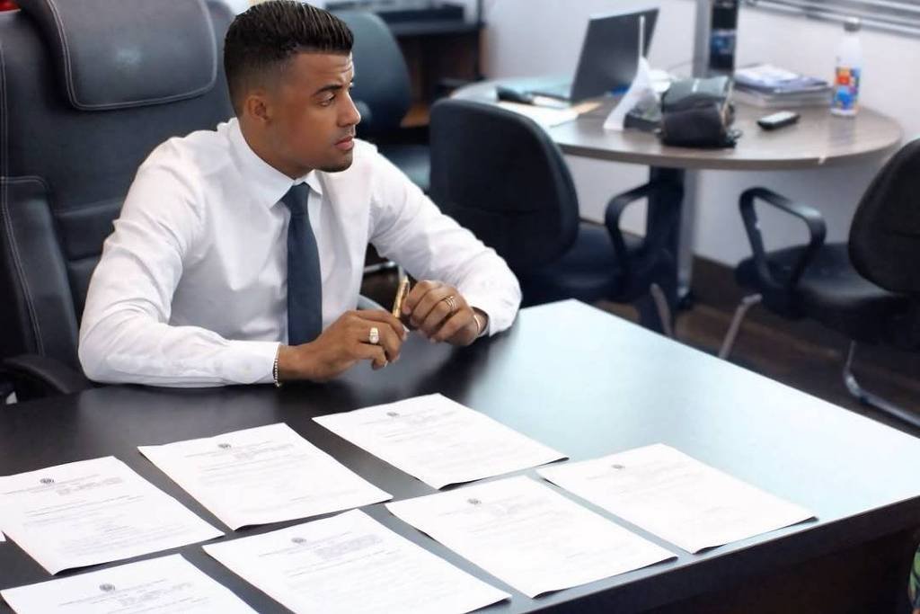 Um homem vestido com camisa branca e gravata está sentado à mesa em um escritório. Ele parece estar concentrado, olhando para documentos dispostos à sua frente. A mesa é escura e há várias folhas de papel organizadas sobre ela. Ao fundo, há cadeiras e uma mesa de escritório, além de janelas que permitem a entrada de luz natural.