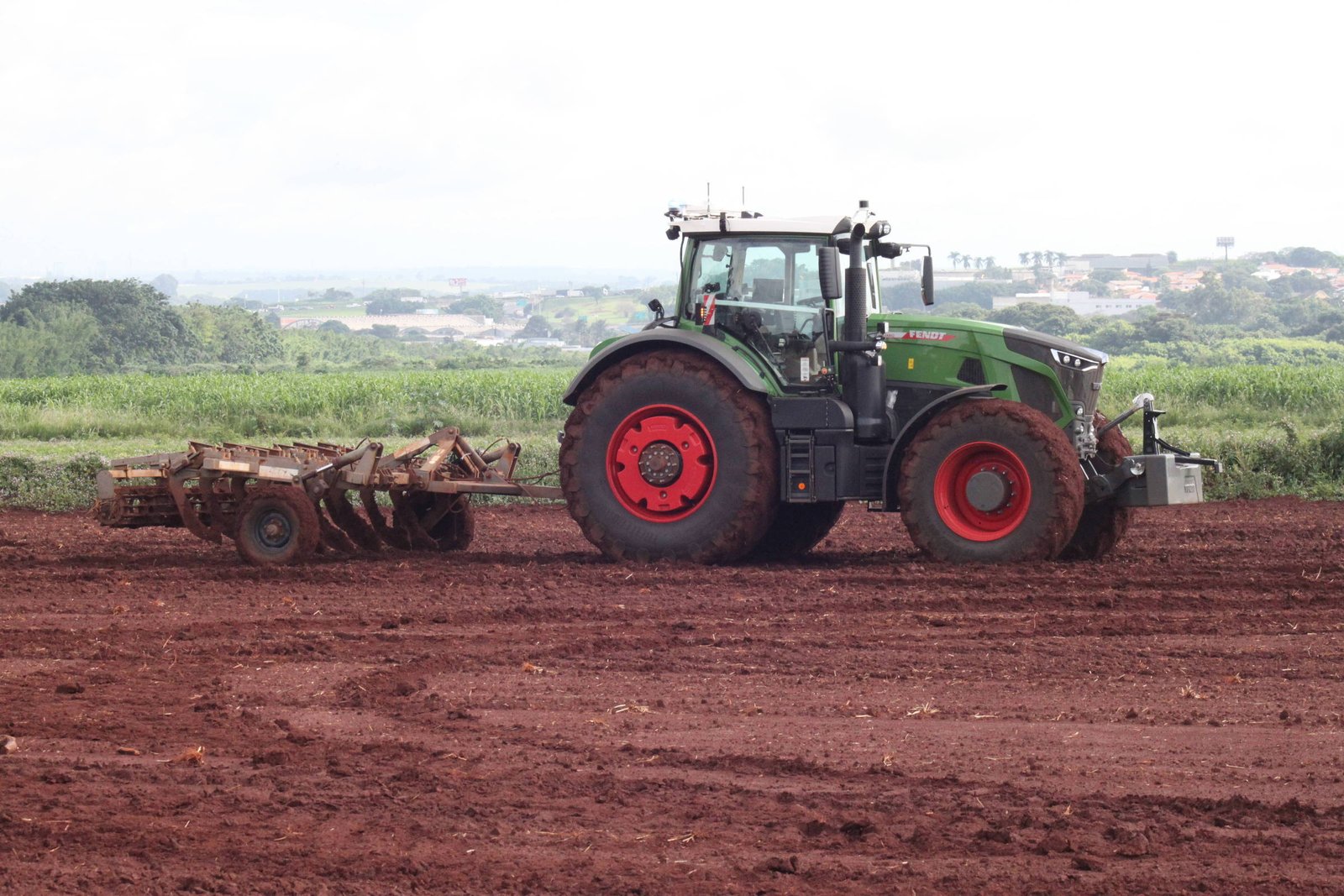Trator verde com rodas vermelhas arando solo vermelho em campo aberto. Vegetação e área urbana ao fundo sob céu nublado.