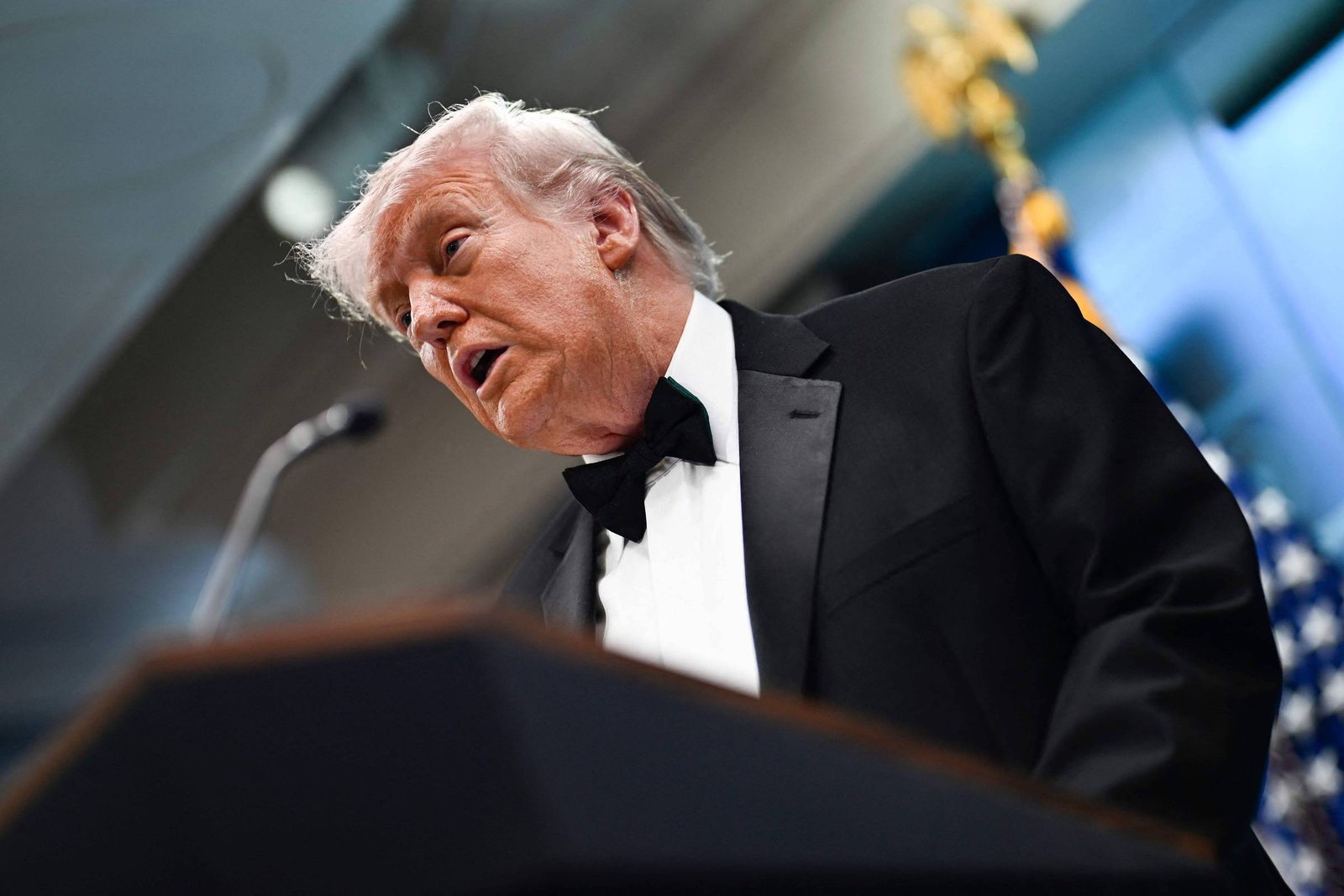 Homem de cabelo branco e terno preto com gravata borboleta fala em púlpito em ambiente interno. Bandeira dos Estados Unidos aparece desfocada ao fundo.