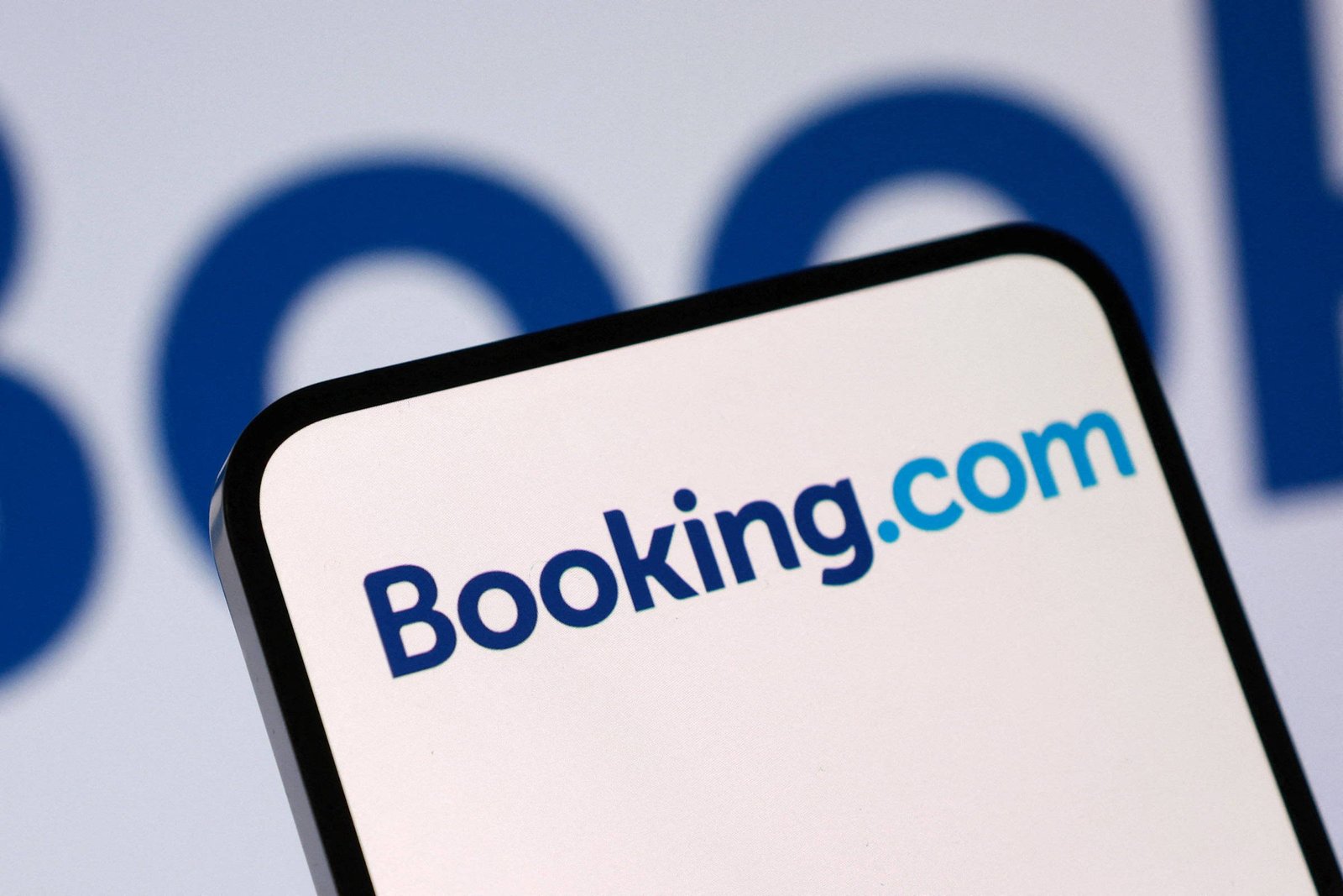 Tela de smartphone mostra o logo do Booking.com em fundo branco, com o texto em azul escuro e azul claro. Ao fundo, desfocado, aparece parte do logo ampliado da mesma marca.