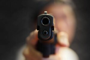 Arma de fogo preta em foco central, apontada diretamente para a lente. Fundo desfocado mostra pessoa segurando a arma, sem detalhes faciais visíveis.