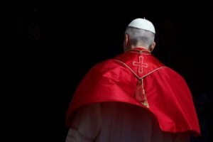 Pessoa vestindo traje religioso branco e capa vermelha com cruz bordada nas costas, vista de costas contra fundo preto